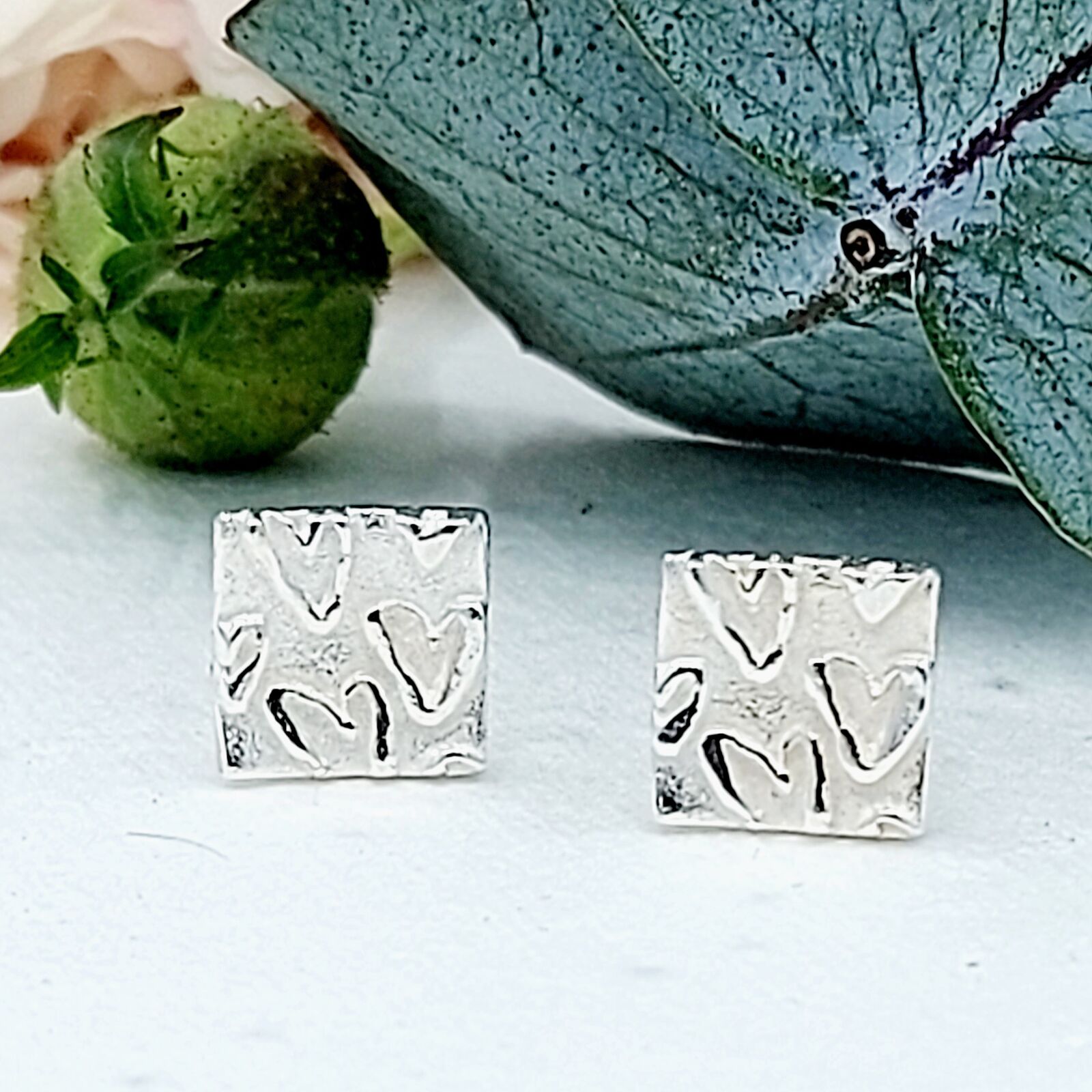 Very cute handcrafted silver square studs with a raised heart pattern that subtly gleams against the frosted matte background.g